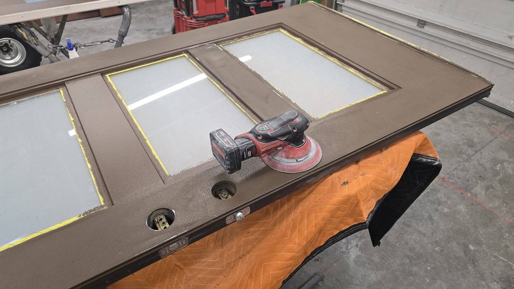 Random-orbit sander resting on the brown entry door, glass lites taped off, in the shop